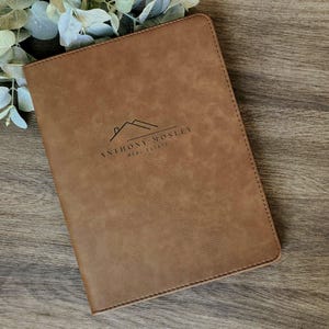 May include: A brown leather-bound notebook with the text "ANTHONY MOSLEY REAL ESTATE" and a house logo. The notebook is rectangular and has a stitched border. The background is a wooden surface with greenery.