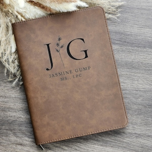 May include: A brown leather journal with the initials "J G" and the name "Jasmine Gump MS., LPC" printed in black. The journal has a textured surface and a silver zipper. Dried flowers are in the background.