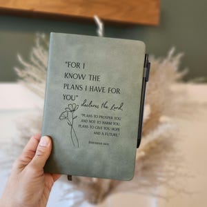 Könnte beinhalten: Ein salbeigrünes Notizbuch mit dem Zitat „DENN ICH KENNE DIE PLÄNE, DIE ICH FÜR EUCH HABE“ und einer floralen Illustration. Das Notizbuch enthält den Text „PLÄNE, EUCH ZU ERMÖGLICHEN UND NICHT ZU SCHADEN, EUCH ZUKUNFT UND HOFFNUNG ZU GEBEN“ und die Referenz Jeremia 29:11. Ein schwarzer Stift ist befestigt.