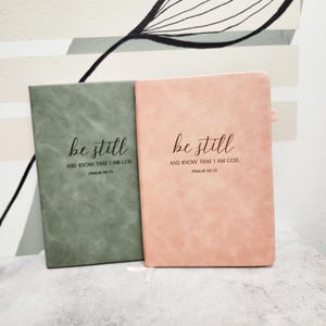 May include: Two journals, one sage green and one dusty rose, are displayed side-by-side. The text "be still AND KNOW THAT I AM GOD. PSALM 46:10" is printed in a delicate script on the front of each journal. The journals have a textured appearance.