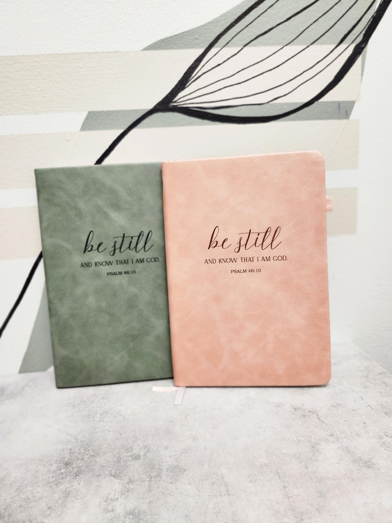 May include: Two journals, one sage green and one dusty rose, are displayed side-by-side. The text "be still AND KNOW THAT I AM GOD. PSALM 46:10" is printed in a delicate script on the front of each journal. The journals have a textured appearance.