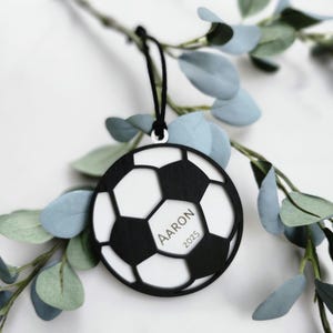 May include: A black and white soccer ball ornament with the name "Aaron" and the year "2025" printed on it. The ornament is hanging from a black string and is surrounded by green and blue leaves.