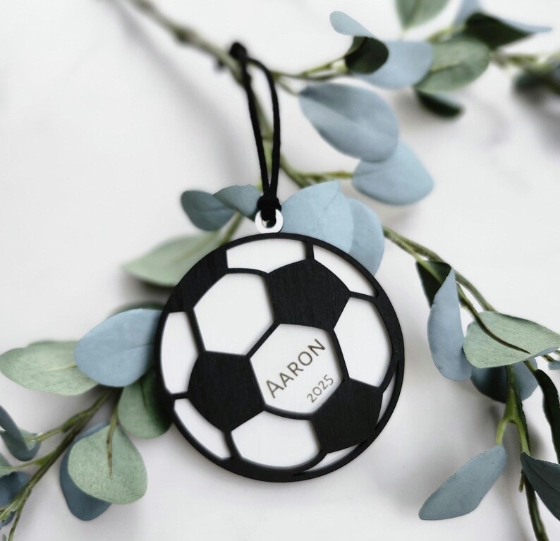 May include: A black and white soccer ball ornament with the name "Aaron" and the year "2025" printed on it. The ornament is hanging from a black string and is surrounded by green and blue leaves.