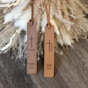 May include: Two leather bookmarks with brown straps. Each bookmark has a cross, the name "CALEB", and the text "CONFIRMED IN CHRIST MAY 2026". The bookmarks are in shades of brown and tan.
