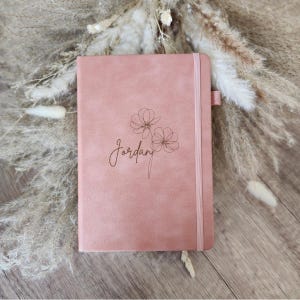 May include: A dusty rose-colored journal with the name "Jordan" and floral design. The journal has an elastic band closure and a ribbon bookmark. The cover is a solid color with a debossed floral design.