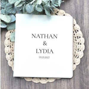 May include: A square white album with a smooth cover featuring the black printed text NATHAN & LYDIA and the date 05.25.2027 centered on the front. The album has stitched edges and a clean, flat surface design for document or photo storage.