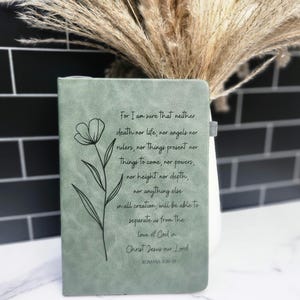 May include: Sage green journal with a floral line drawing and the text of Romans 8:38-39. The journal has a simple, elegant design, perfect for writing or note-taking. The cover features a minimalist flower illustration.