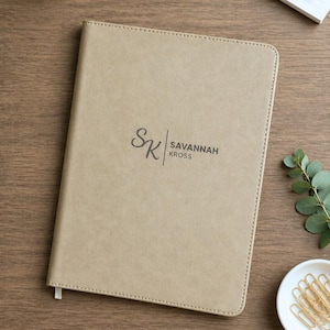 May include: Tan leather-bound journal with the initials "SK" and the name "Savannah Kross" printed in dark gray. The journal has a stitched border and a ribbon bookmark. The journal is on a wooden surface.