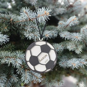 May include: A black and white soccer ball ornament with the name "Aaron" and the year "2025" engraved on it. The ornament is hanging from a black string against a backdrop of a blue-green evergreen tree.