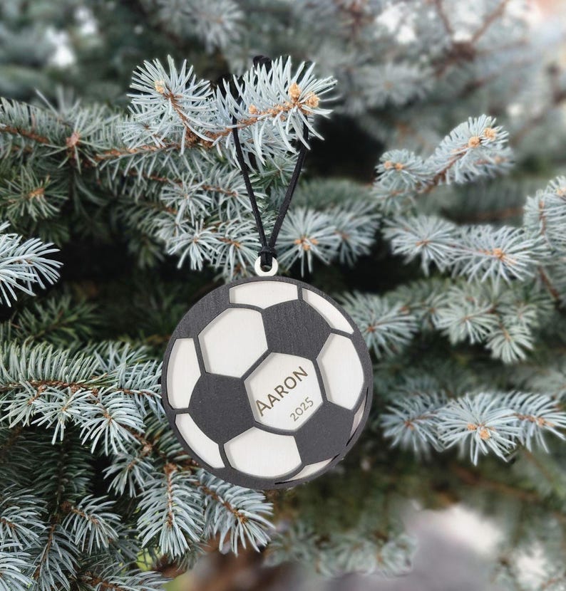 May include: A black and white soccer ball ornament with the name "Aaron" and the year "2025" engraved on it. The ornament is hanging from a black string against a backdrop of a blue-green evergreen tree.