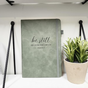 May include: Sage green journal with the words "be still and know that I am God" printed on the cover. The journal has a gray elastic band closure and a ribbon bookmark. A small potted plant sits to the right.