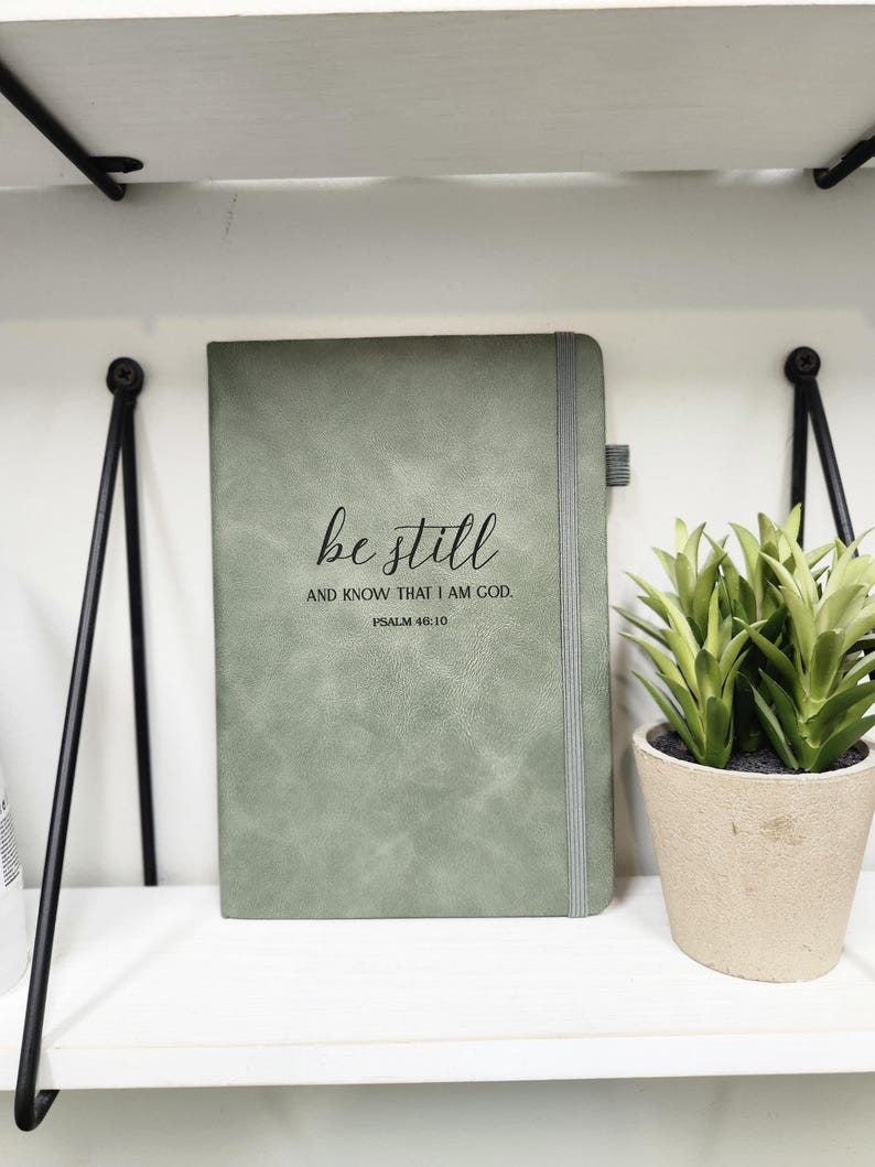 May include: Sage green journal with the words "be still and know that I am God" printed on the cover. The journal has a gray elastic band closure and a ribbon bookmark. A small potted plant sits to the right.