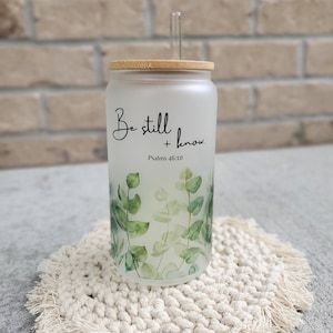 May include: A frosted glass tumbler with a bamboo lid and a clear straw. The tumbler has a green leafy design and the text "Be still and know. Psalms 46:10" printed on it. The tumbler is sitting on a white crocheted coaster.