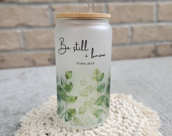 May include: A frosted glass tumbler with a bamboo lid and a clear straw. The tumbler has a green leafy design and the text "Be still and know. Psalms 46:10" printed on it. The tumbler is sitting on a white crocheted coaster.