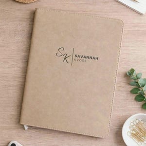 May include: Tan-colored journal with the text "SK | SAVANNAH KROSS" in black. The journal has a smooth, leather-like texture and a white ribbon bookmark. The journal is on a light-colored wooden surface.