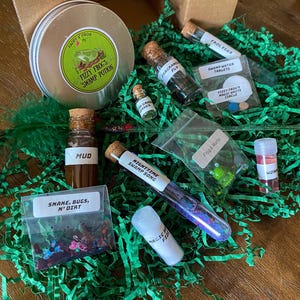 May include: A whimsical swamp-themed kit with various potion ingredients. Includes a tin labeled "Fizzy Frog's Swamp Potion," vials of "Mud," "Fragrant Fly," and "Nighttime Swamp Song," and packets of "Snake, Bugs, N' Dirt" and "Frog Eggs."