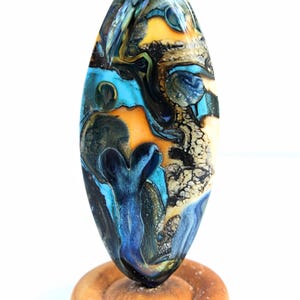 May include: An oval-shaped glass bead featuring a marbled pattern of turquoise, deep blue, black, and golden yellow colors. The bead has a glossy, smooth surface and is displayed upright on a small, circular, light-colored wooden base.