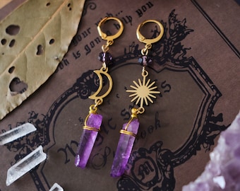 Puede incluir: Un par de pendientes de oro con gotas de cristal de amatista, un amuleto de luna creciente y un amuleto de sol. Los pendientes se muestran en un libro con el texto "Precious Linenote, Idea, Since" y un cúmulo de cristal de amatista púrpura.