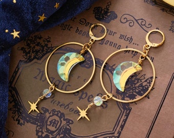 May include: A pair of gold hoop earrings with crescent moon and star charms. The earrings are on a brown book with gold lettering and a purple amethyst crystal.
