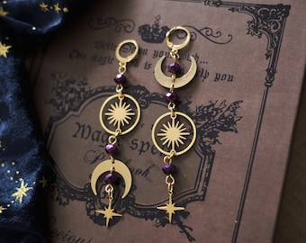 May include: A pair of gold-toned earrings with purple glass beads, crescent moons, and star charms. The earrings are hanging from a book with the text "Magic Spellbook" on the cover.