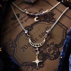 Può includere: Una collana d'argento con un ciondolo a forma di mezzaluna e un ciondolo a stella, ornata da perline viola. La collana è sovrapposta a un libro vintage con il testo "Magic Spells". È visibile una seconda collana con un ciondolo a forma di mezzaluna.