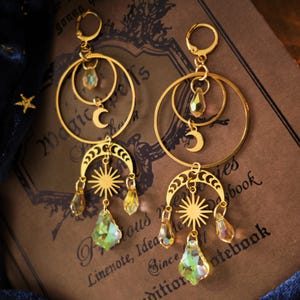 Puede incluir: Pendientes dorados con motivos celestiales, sol y luna. Cada pendiente tiene múltiples aros, una luna creciente, un sol y colgantes de cristal iridiscente. Los pendientes están sobre un cuaderno vintage y tela azul oscuro con estrellas doradas. El texto dice 'Linenote, Idea'.