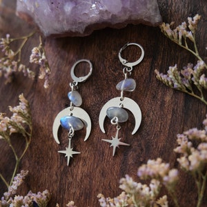 Puede incluir: Pendientes de plata con colgantes de luna y estrella y cuentas de piedra lunar iridiscente. Los pendientes se muestran sobre una superficie de madera con amatista y flores secas.