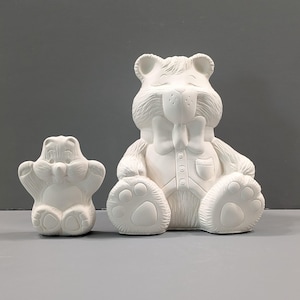 May include: Two unpainted ceramic bear figurines. The larger bear is seated, wearing a bow tie and vest. The smaller bear is standing with its arms raised. Both have detailed paw prints and facial features, ready for painting.