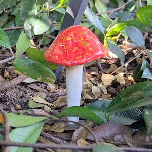 May include: A ceramic mushroom sculpture with a vibrant red cap speckled with yellow and green spots. The stem is white, and the mushroom is surrounded by green leaves and brown foliage, creating a natural garden aesthetic.