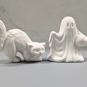 May include: Two unpainted white ceramic figurines. One figurine is a cat in a crouching position with a bushy tail. The second figurine is a ghost draped in a sheet, holding a small round pumpkin shape at its side. Both items have a matte finish.
