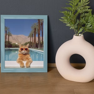 May include: A framed photograph of a cat wearing sunglasses and sitting by a pool. The cat is orange and white and the pool is surrounded by palm trees.