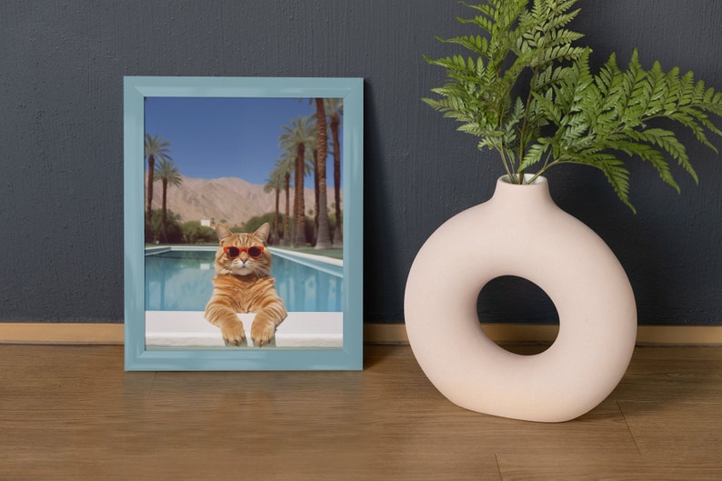 May include: A framed photograph of a cat wearing sunglasses and sitting by a pool. The cat is orange and white and the pool is surrounded by palm trees.