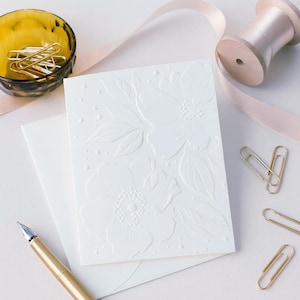 May include: A white embossed floral greeting card with an envelope, a gold pen, and gold paper clips. A spool of pink ribbon and a small amber glass bowl with gold paper clips are also present. The card features a detailed floral design.