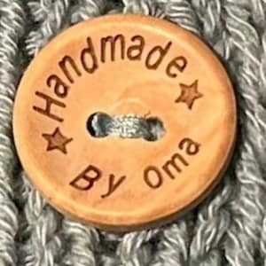 Könnte beinhalten: Nahaufnahme eines hellbraunen Knopfes mit dem Text "Handmade by Oma" und zwei Sternen auf einer grauen gehäkelten Oberfläche.