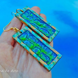 May include: A pair of rectangular earrings with a green and blue iridescent finish. The earrings have a gold-colored border and a gold-colored hook.