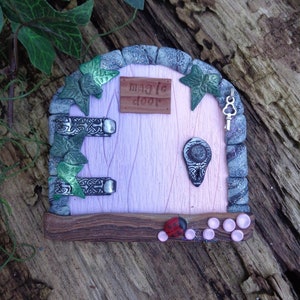 Puede incluir: Una puerta de hada rosa en miniatura con un letrero de madera marrón que dice "magic door" y una cerradura plateada. La puerta está decorada con hiedra verde y detalles plateados. Hay una mariquita y puntos rosas en la parte inferior de la puerta.