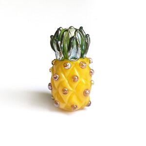 Könnte beinhalten: Eine gelbe Glasperle in Form einer Ananas mit grünem, blattförmigem Oberteil und violetten Akzenten. Die Perle ist eine realistische Darstellung einer Ananas.