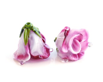 Könnte beinhalten: Zwei rosafarbene Glasrosenperlen mit weißen Zentren und grünen Blättern. Die Perlen sind so gestaltet, dass sie wie Rosen aussehen.