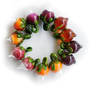May include: A circular arrangement of colorful glass fruit sculptures. The collection includes pieces in shades of red, orange, and purple, each with green stems and white tips. The glass fruit is displayed against a white background.