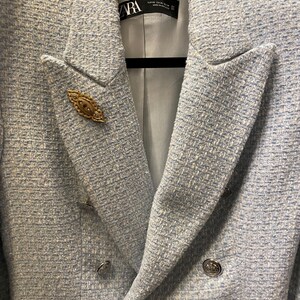 May include: A light blue blazer with a double breasted closure and silver buttons. The blazer has a textured fabric and a gold pin with a red stone in the center.