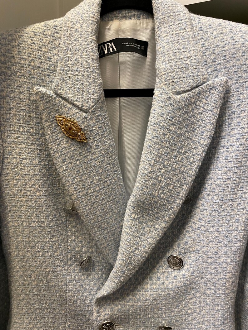 May include: A light blue blazer with a double breasted closure and silver buttons. The blazer has a textured fabric and a gold pin with a red stone in the center.