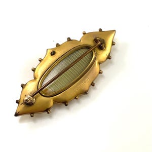 May include: A gold-toned brooch with a decorative design and a glass or plastic oval insert. The back of the brooch has a marking that reads "B.D. & S.O."