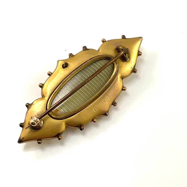 May include: A gold-toned brooch with a decorative design and a glass or plastic oval insert. The back of the brooch has a marking that reads "B.D. & S.O."
