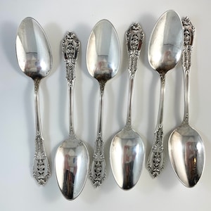 May include: Six silver teaspoons with ornate handles. The spoons have a polished finish and are arranged in a row.