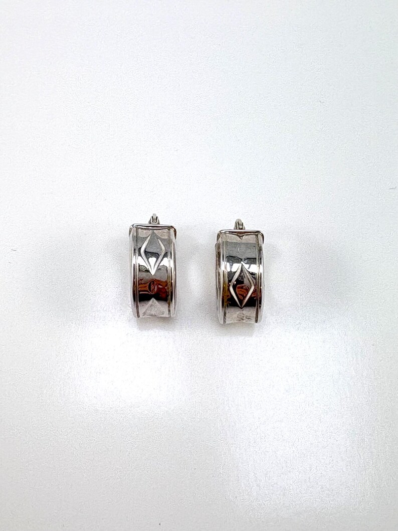 Puede incluir: Un par de pendientes de aro en tono plateado con acabado pulido y un motivo central grabado en forma de diamante. Los pendientes tienen un dise&ntilde;o de banda ancha y curva con un mecanismo de cierre de bisagra visible en la parte superior.