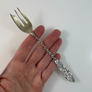 May include: A silver fork with a twisted handle and a floral design on the end. The fork is approximately 15 cm long.
