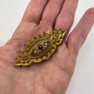 May include: A gold brooch with an oval center featuring a red stone surrounded by two white pearls. The brooch is decorated with intricate floral designs.