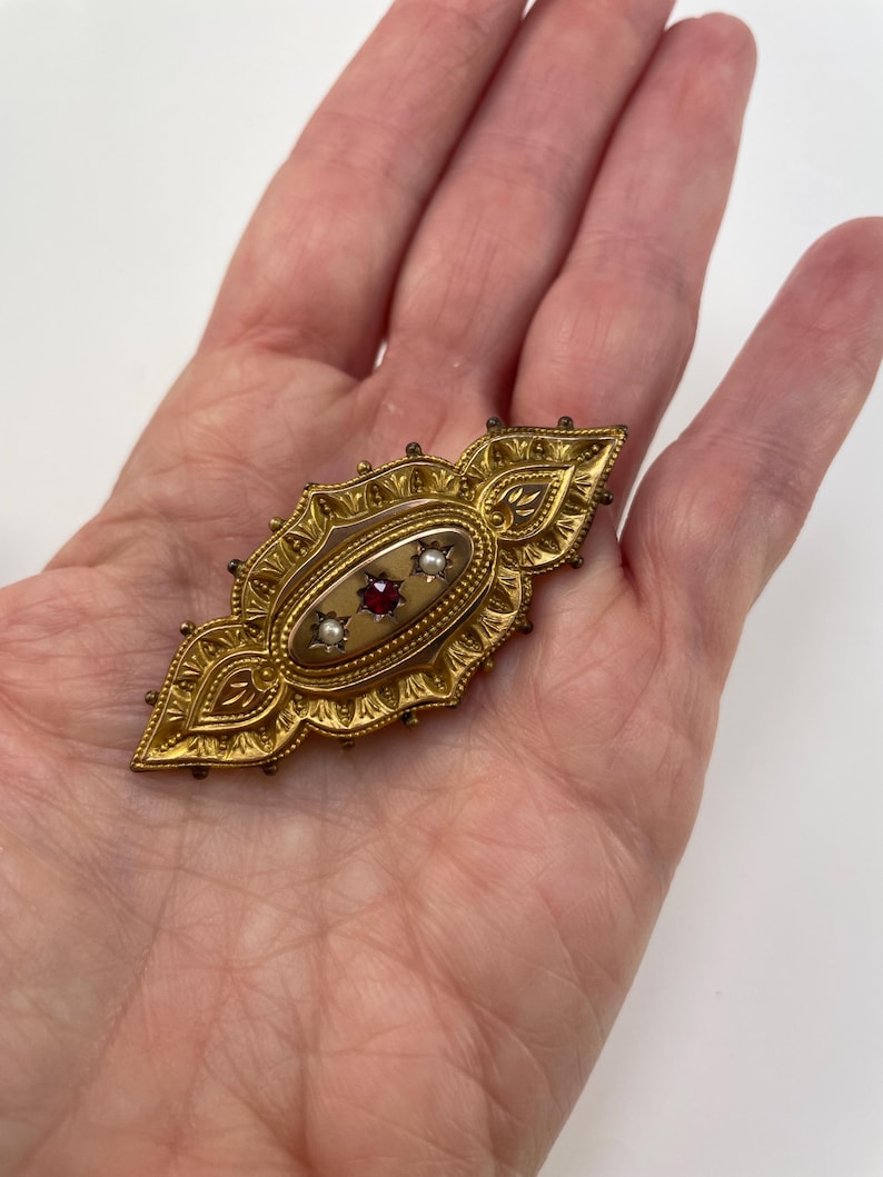 May include: A gold brooch with an oval center featuring a red stone surrounded by two white pearls. The brooch is decorated with intricate floral designs.