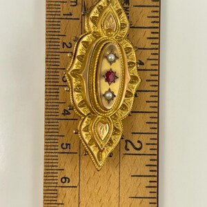 May include: A gold-toned brooch with a red and white stone design. The brooch is about 2 inches long and is measured against a ruler.