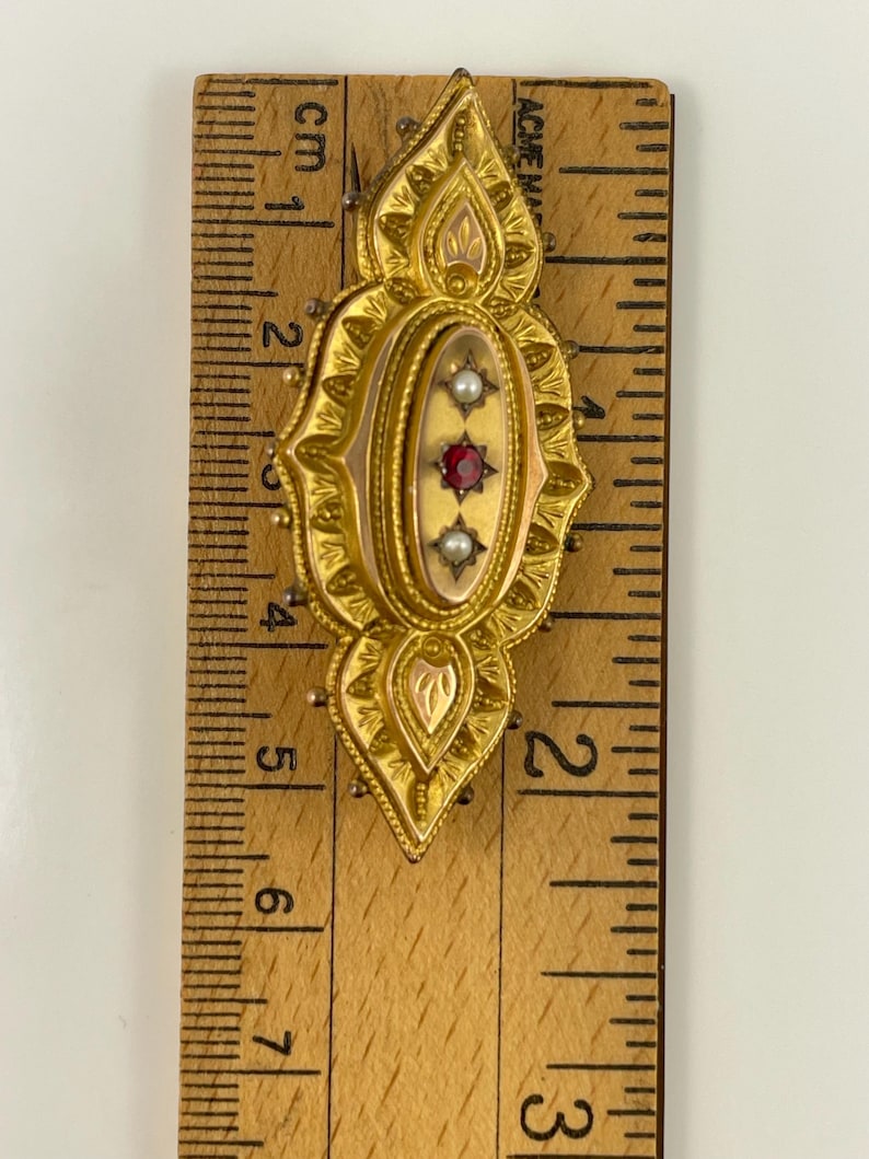 May include: A gold-toned brooch with a red and white stone design. The brooch is about 2 inches long and is measured against a ruler.